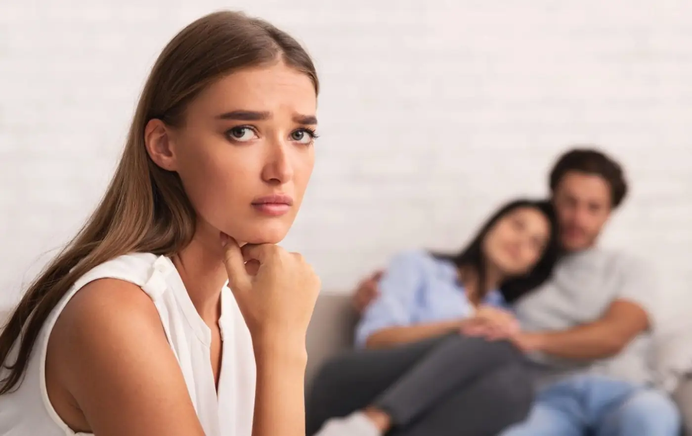 Junge Frau im Vordergrund und ihr Ex mit einer anderen Frau auf dem Sofa im Hintergrund. Symbolbild für Ex-Freundin eifersüchtig machen.